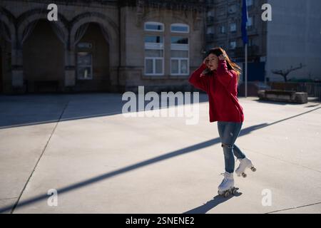 Giovane donna che indossa un maglione rosso vivace e un elegante pattinaggio a rotelle in una vivace piazza della città, immersa nel caldo sole e si diverte con ener Foto Stock