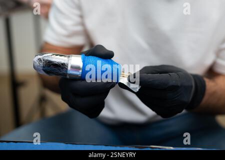 Tatuatore che indossa guanti neri per pulire e preparare accuratamente una macchina per tatuaggi avvolta in plastica e nastro blu, garantendo igiene e precisione Foto Stock