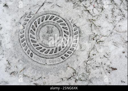 Un primo piano di un tombino parzialmente coperto da uno strato sottile di neve, rivelando il suo schema geometrico circolare. L'impronta della copertina contrasta Foto Stock