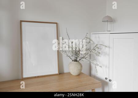 Natura morta degli interni scandinavi in primavera. Mockup con cornice in legno sottile. Rami di prugna ciliegina bianca in fiore in vaso di ceramica su bambù Foto Stock