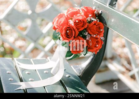 Un vivace bouquet di rose rosse con nastri si trova graziosamente su una panchina, circondato da colorate foglie autunnali. Foto Stock