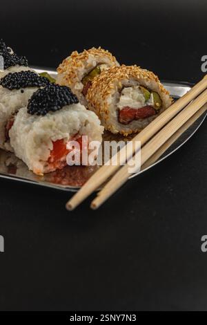 Gli involtini di sushi con varie ripieni sono disposti in modo ordinato su un piatto d'argento lucido. Due set di bacchette sono posizionati lateralmente, per aggiungere un tocco elegante Foto Stock