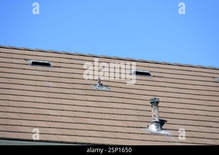 Vista ravvicinata di un tetto inclinato con piastrelle marroni, con due aperture di ventilazione e un tubo di camino in metallo intemprato sotto un cielo azzurro Foto Stock