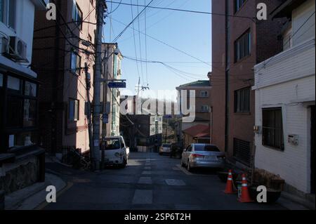Vicolo stretto, Corea del Sud. Auto parcheggiate su entrambi i lati. Gli edifici su entrambi i lati gettano lunghe ombre, un senso di tranquilla solitudine. L'immagine evoca una fe Foto Stock