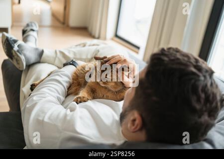 Vista dall'alto dell'uomo che si rilassa sul divano con un gatto marrone sul petto Foto Stock