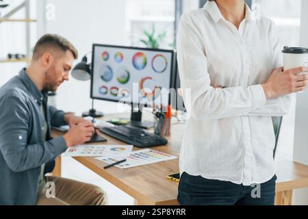 Donna in piedi davanti a un uomo che sedeva accanto al tavolo. Due designer che lavorano insieme in ufficio. Foto Stock