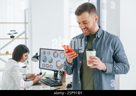 Uomo positivo in piedi davanti a una donna seduta accanto al tavolo. Due designer che lavorano insieme in ufficio. Foto Stock