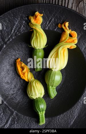 Zucchine fresche giovani con fiori in piatto nero. Vista dall'alto dei fiori di zucchine fresche. Fotografia gastronomica Foto Stock