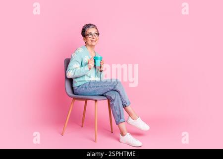 Donna più anziana allegra in un casual maglione floreale seduto su una sedia con sfondo rosa, tenendo in mano una tazza Foto Stock