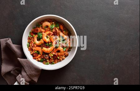 Deliziosa jambalaya creola con gamberi, salsicce affumicate e verdure in padella chiara su sfondo marrone scuro. Con spazio libero. Vista dall'alto, disposizione piatta Foto Stock