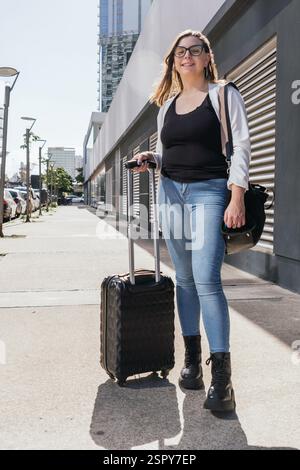 Una giovane donna che indossa occhiali, t-shirt nera, giacca leggera e jeans cammina con valigia e borsa nera sulla strada urbana vicino a edifici moderni, divertiti Foto Stock