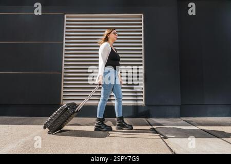 Giovane donna d'affari che tira i bagagli su un marciapiede soleggiato della città, passeggia davanti a un elegante edificio moderno, incarnando l'essenza del viaggio urbano Foto Stock