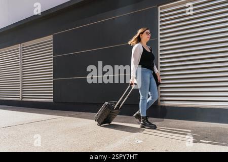 Donna d'affari che tira la valigia del trolley mentre cammina su un marciapiede in città durante una giornata di sole, vicino a un edificio moderno con facciata metallica Foto Stock