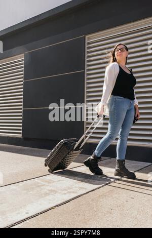 Giovane donna d'affari che tira la valigia mentre cammina fuori dal terminal dell'aeroporto, si prepara a imbarcarsi in un viaggio di lavoro, rappresentando viaggi, autobus Foto Stock