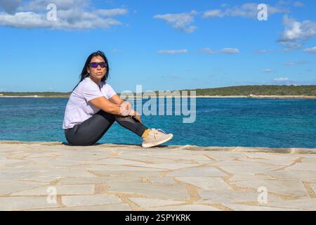 Donna seduta accanto all'oceano, che appare serena, bella latina sul mare Foto Stock