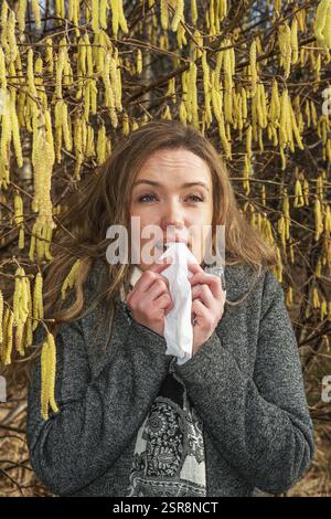 La giovane donna caucasica con problemi di allergia si trova in una fioritura di nocciole e starnutisce in un fazzoletto a Ystad, contea di Skane, Svezia, Scandinavia Foto Stock
