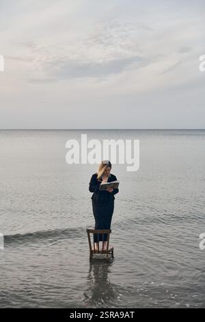 Una giovane donna adulta sta su una sedia di legno nelle calme acque della spiaggia, leggendo un libro. Il cielo sereno e le onde dolci creano un'atmosfera tranquilla ma surreale, simbo Foto Stock