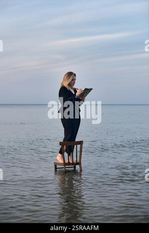 Una giovane donna adulta sta su una sedia di legno nelle calme acque della spiaggia, leggendo un libro. Il cielo sereno e le onde dolci creano un'atmosfera tranquilla ma surreale, simbo Foto Stock