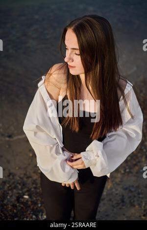 La giovane donna sta serenamente sulla spiaggia rocciosa, mentre la sua camicetta bianca cattura la luce soffusa mentre chiude gli occhi. La scena evoca un umore di tranquilla riflessione e pisello Foto Stock