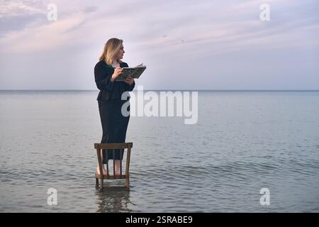 Una giovane donna adulta sta su una sedia di legno nelle calme acque della spiaggia, leggendo un libro. Il cielo sereno e le onde dolci creano un'atmosfera tranquilla ma surreale, simbo Foto Stock