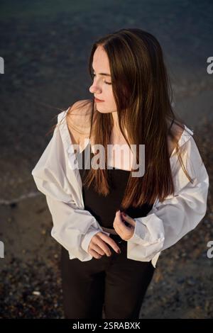 La giovane donna sta serenamente sulla spiaggia rocciosa, mentre la sua camicetta bianca cattura la luce soffusa mentre chiude gli occhi. La scena evoca un umore di tranquilla riflessione e pisello Foto Stock