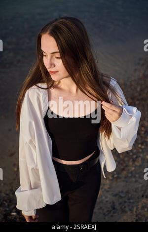 La giovane donna sta serenamente sulla spiaggia rocciosa, mentre la sua camicetta bianca cattura la luce soffusa mentre chiude gli occhi. La scena evoca un umore di tranquilla riflessione e pisello Foto Stock