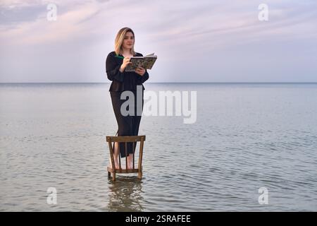 Una giovane donna adulta sta su una sedia di legno nelle calme acque della spiaggia, leggendo un libro. Il cielo sereno e le onde dolci creano un'atmosfera tranquilla ma surreale, simbo Foto Stock