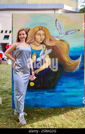 Giovane artista donna con i capelli castani si erge orgogliosamente di fronte a colorate sirene. La luce del giorno migliora i colori vivaci. Circondata dal verde, sorride Foto Stock