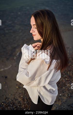 La giovane donna sta serenamente sulla spiaggia rocciosa, mentre la sua camicetta bianca cattura la luce soffusa mentre chiude gli occhi. La scena evoca un umore di tranquilla riflessione e pisello Foto Stock