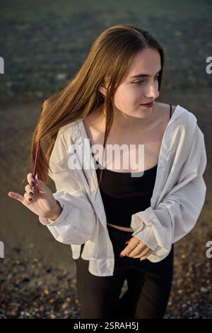 Una giovane donna si staglia serenamente lungo il litorale, drappeggiata in camicia bianca su un outfit nero. Il sole tramonta diffonde una calda luce soffusa, migliorando la calma e l'atmosfera riflessiva di Foto Stock