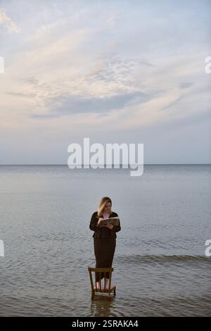 Una giovane donna adulta sta su una sedia di legno nelle calme acque della spiaggia, leggendo un libro. Il cielo sereno e le onde dolci creano un'atmosfera tranquilla ma surreale, simbo Foto Stock