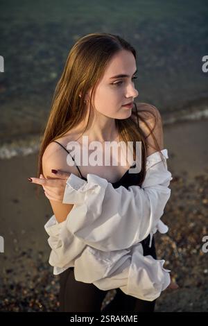 Una giovane donna si staglia serenamente lungo il litorale, drappeggiata in camicia bianca su un outfit nero. Il sole tramonta diffonde una calda luce soffusa, migliorando la calma e l'atmosfera riflessiva di Foto Stock