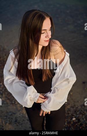 La giovane donna sta serenamente sulla spiaggia rocciosa, mentre la sua camicetta bianca cattura la luce soffusa mentre chiude gli occhi. La scena evoca un umore di tranquilla riflessione e pisello Foto Stock