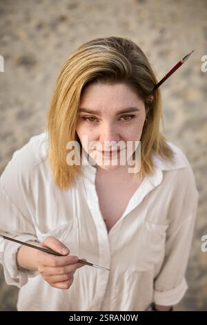 Una giovane donna si erge sulla spiaggia di sabbia, illuminata delicatamente dalla luce naturale. Ha un pennello nei capelli, trasudando creatività e tranquillità. Il suo vestito bianco enh Foto Stock