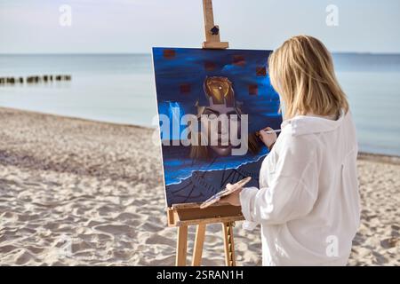 Giovane artista che dipinge ritratti mistici su tela in spiaggia tranquilla. L'opera raffigura un volto femminile surreale circondato da elementi astratti, evocativi Foto Stock