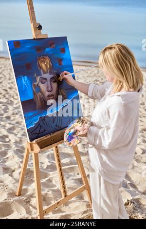 Giovane artista che dipinge ritratti mistici su tela in spiaggia tranquilla. L'opera raffigura un volto femminile surreale circondato da elementi astratti, evocativi Foto Stock