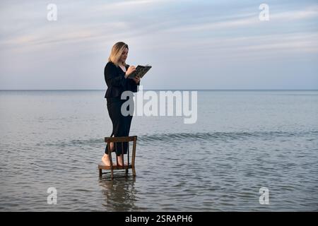 Una giovane donna adulta sta su una sedia di legno nelle calme acque della spiaggia, leggendo un libro. Il cielo sereno e le onde dolci creano un'atmosfera tranquilla ma surreale, simbo Foto Stock