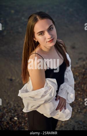 Una giovane donna si staglia serenamente lungo il litorale, drappeggiata in camicia bianca su un outfit nero. Il sole tramonta diffonde una calda luce soffusa, migliorando la calma e l'atmosfera riflessiva di Foto Stock