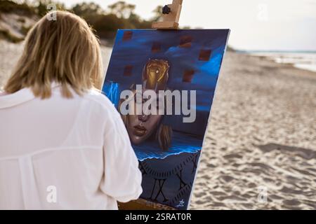 Giovane artista che dipinge ritratti mistici su tela in spiaggia tranquilla. L'opera raffigura un volto femminile surreale circondato da elementi astratti, evocativi Foto Stock
