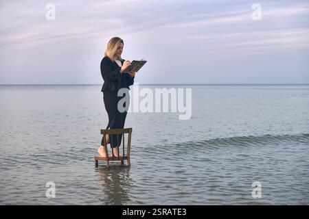 Una giovane donna adulta sta su una sedia di legno nelle calme acque della spiaggia, leggendo un libro. Il cielo sereno e le onde dolci creano un'atmosfera tranquilla ma surreale, simbo Foto Stock