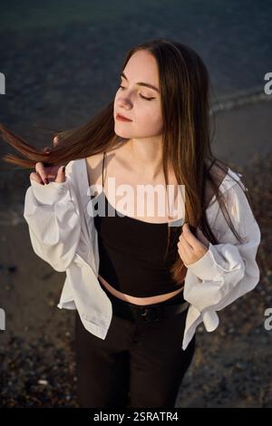 La giovane donna sta serenamente sulla spiaggia rocciosa, mentre la sua camicetta bianca cattura la luce soffusa mentre chiude gli occhi. La scena evoca un umore di tranquilla riflessione e pisello Foto Stock