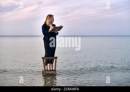 Una giovane donna adulta sta su una sedia di legno nelle calme acque della spiaggia, leggendo un libro. Il cielo sereno e le onde dolci creano un'atmosfera tranquilla ma surreale, simbo Foto Stock