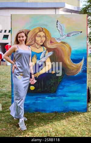 Giovane artista donna con i capelli castani si erge orgogliosamente di fronte a colorate sirene. La luce del giorno migliora i colori vivaci. Circondata dal verde, sorride Foto Stock
