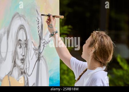 Giovane artista caucasica che dipinge grandi tele nel parco illuminato dal sole. Lo sfondo vibrante mette in risalto la creatività e la concentrazione artistica con colori vivaci Foto Stock