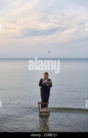 Una giovane donna adulta sta su una sedia di legno nelle calme acque della spiaggia, leggendo un libro. Il cielo sereno e le onde dolci creano un'atmosfera tranquilla ma surreale, simbo Foto Stock