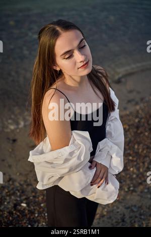 Una giovane donna si staglia serenamente lungo il litorale, drappeggiata in camicia bianca su un outfit nero. Il sole tramonta diffonde una calda luce soffusa, migliorando la calma e l'atmosfera riflessiva di Foto Stock