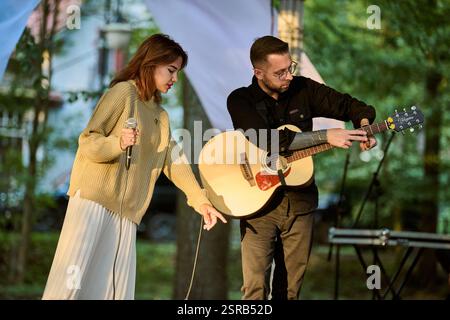 Kaliningrad, Russia - 10.08.2024 - giovane donna canta accanto al chitarrista maschile nel parco illuminato dal sole. Alberi e luce soffusa creano un'atmosfera serena Foto Stock