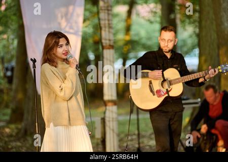 Kaliningrad, Russia - 10.08.2024 - giovane donna canta accanto al chitarrista maschile nel parco illuminato dal sole. Alberi e luce soffusa creano un'atmosfera serena Foto Stock