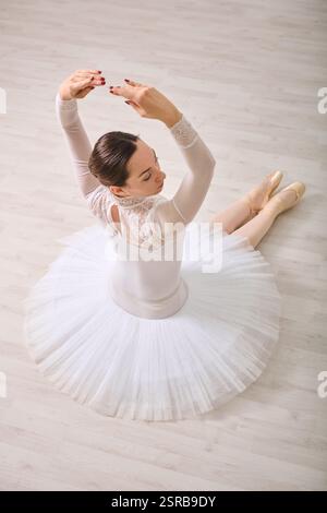 Giovane ballerina femminile con capelli scuri in tutù bianco classico seduta su pavimento in legno, braccia sollevate elegantemente. L'illuminazione soffusa mette in risalto la postura aggraziata Foto Stock