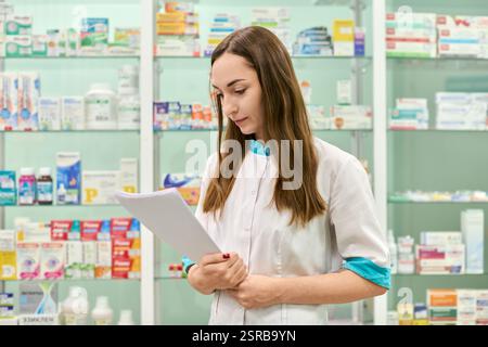 Giovane donna farmacista adulta con capelli castani, caucasico, revisione prescrizione in farmacia illuminata. Scaffali di medicinali e prodotti in backgr Foto Stock
